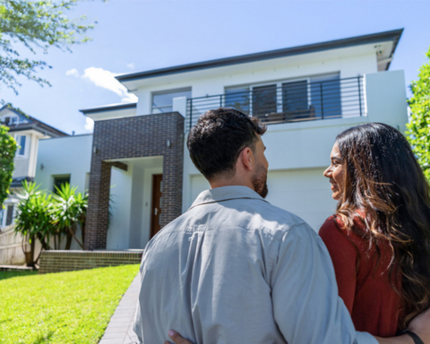A young couple standing in the driveway of a suburban home smiling at each other