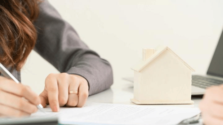 Woman sitting at desk writing on paper, with a small wooden house and laptop in front of her