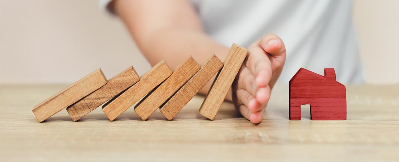 A row of wooden dominoes tilted towards a red house block, with a hand dividing them to prevent the dominoes from crashing into the house
