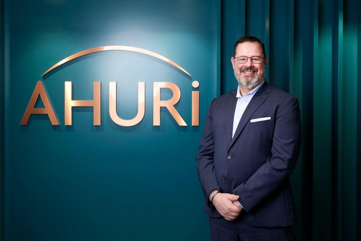 Dr Michael Fotheringham standing beside the AHURI logo in their Melbourne office. Michael is wearing a suit with no tie and is smiling at the camera. The logo is copper lettering against a teal coloured wall.