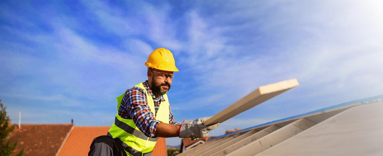 A housing construction worker standing on roof in hard hat and protective gear as he places plank down.