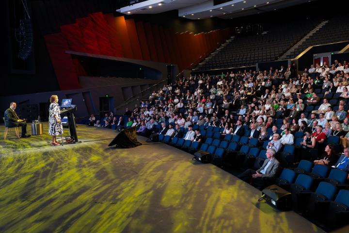 Welcome to Country at NHC25. Side view of speaker on stage and large auditorium full of people.