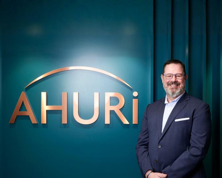 Dr Michael Fotheringham standing beside the AHURI logo at their Melbourne office. Michael is wearing a suit with no tie and smiling at the camera. The logo is in copper lettering against a teal coloured wall.