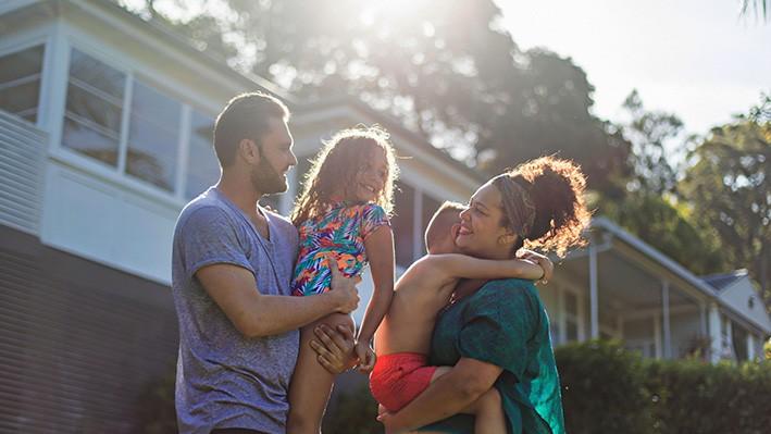Stock image of an Australian family in the garden at home on a sunny day. The mum is holding her son, approx 6 years old, and the dad is holding his daughter, approx 8 years old.