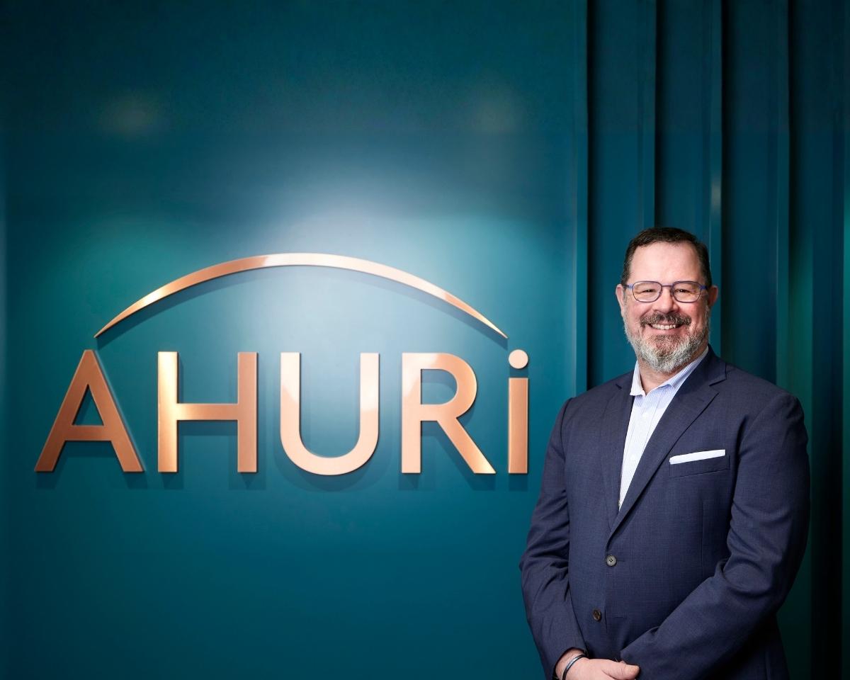Dr Michael Fotheringham standing beside the AHURI logo at their Melbourne office. Michael is wearing a suit with no tie and smiling at the camera. The logo is in copper lettering against a teal coloured wall.