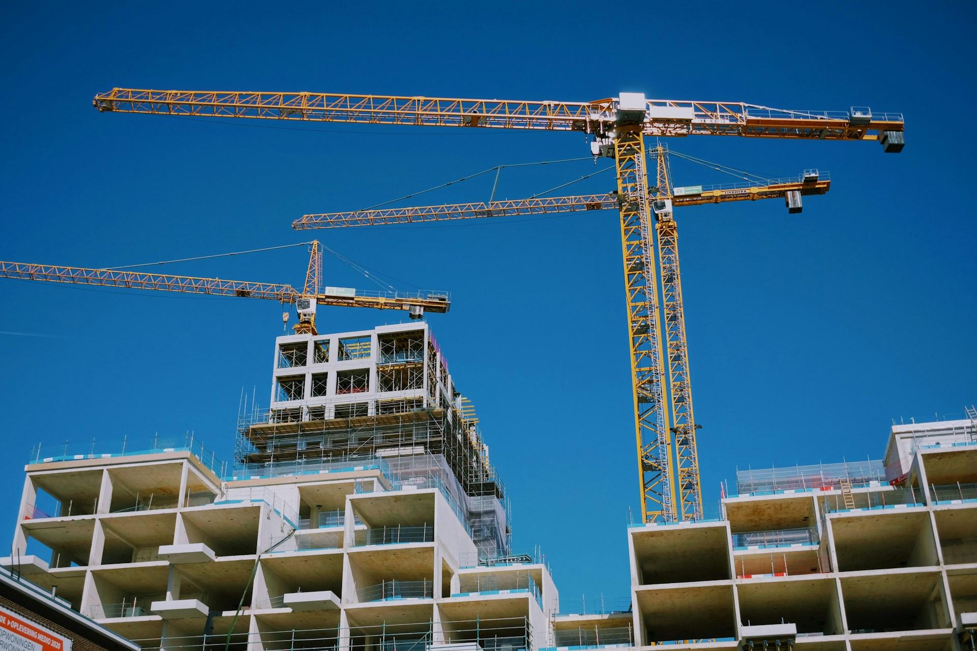 Large cranes above apartment construction project against bright blue sky. By @marekminor on Unsplash.